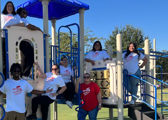7 women and 1 man standing around a playground set with white t shirts. One lady is wearing a red tshirt.